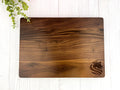 A rectangular walnut cutting board with a distinctive grain and a engraved logo near the bottom corner, placed on a white surface with a partial view of a green plant to the left.
