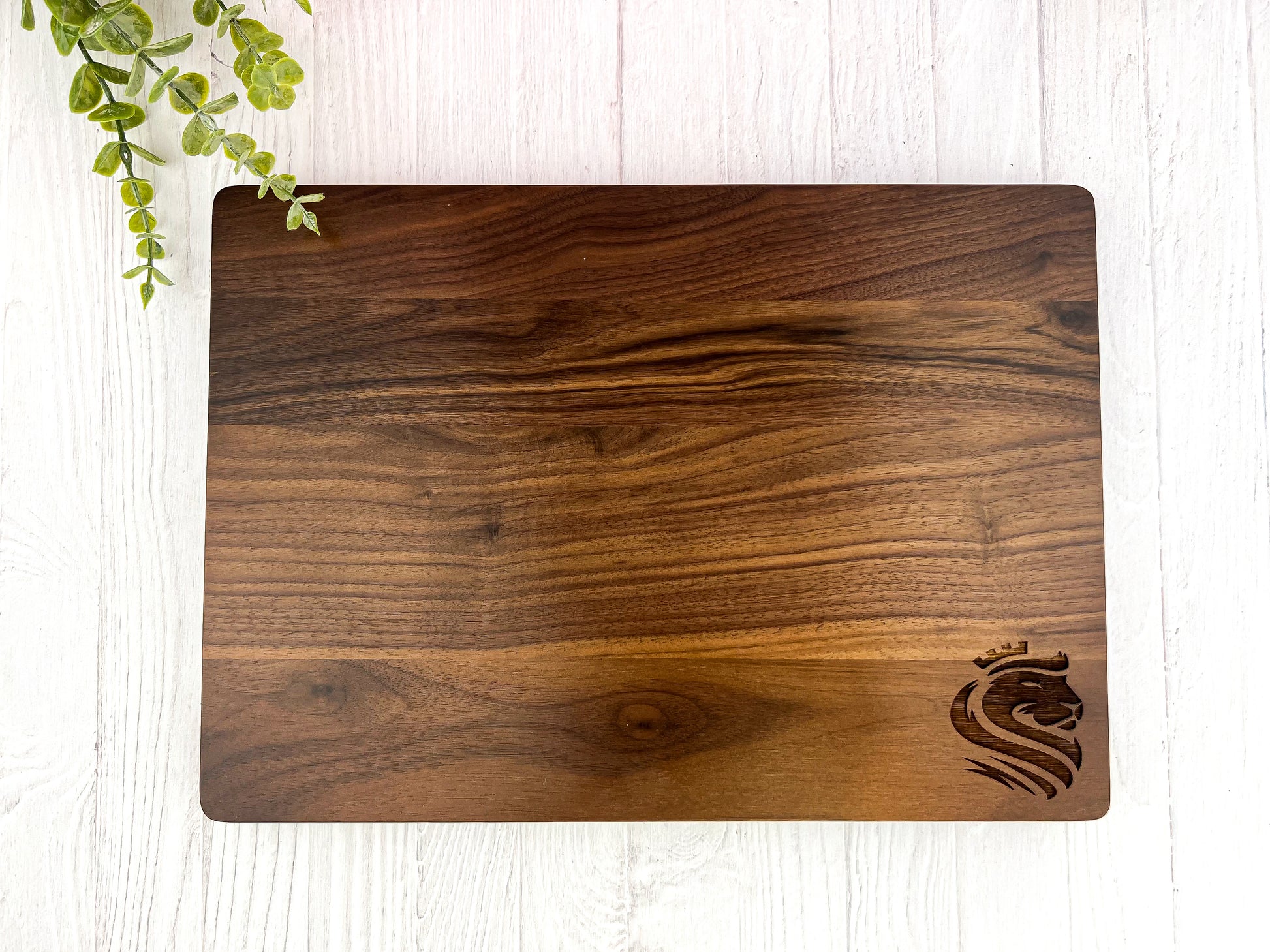 A rectangular walnut cutting board with a distinctive grain and a engraved logo near the bottom corner, placed on a white surface with a partial view of a green plant to the left.