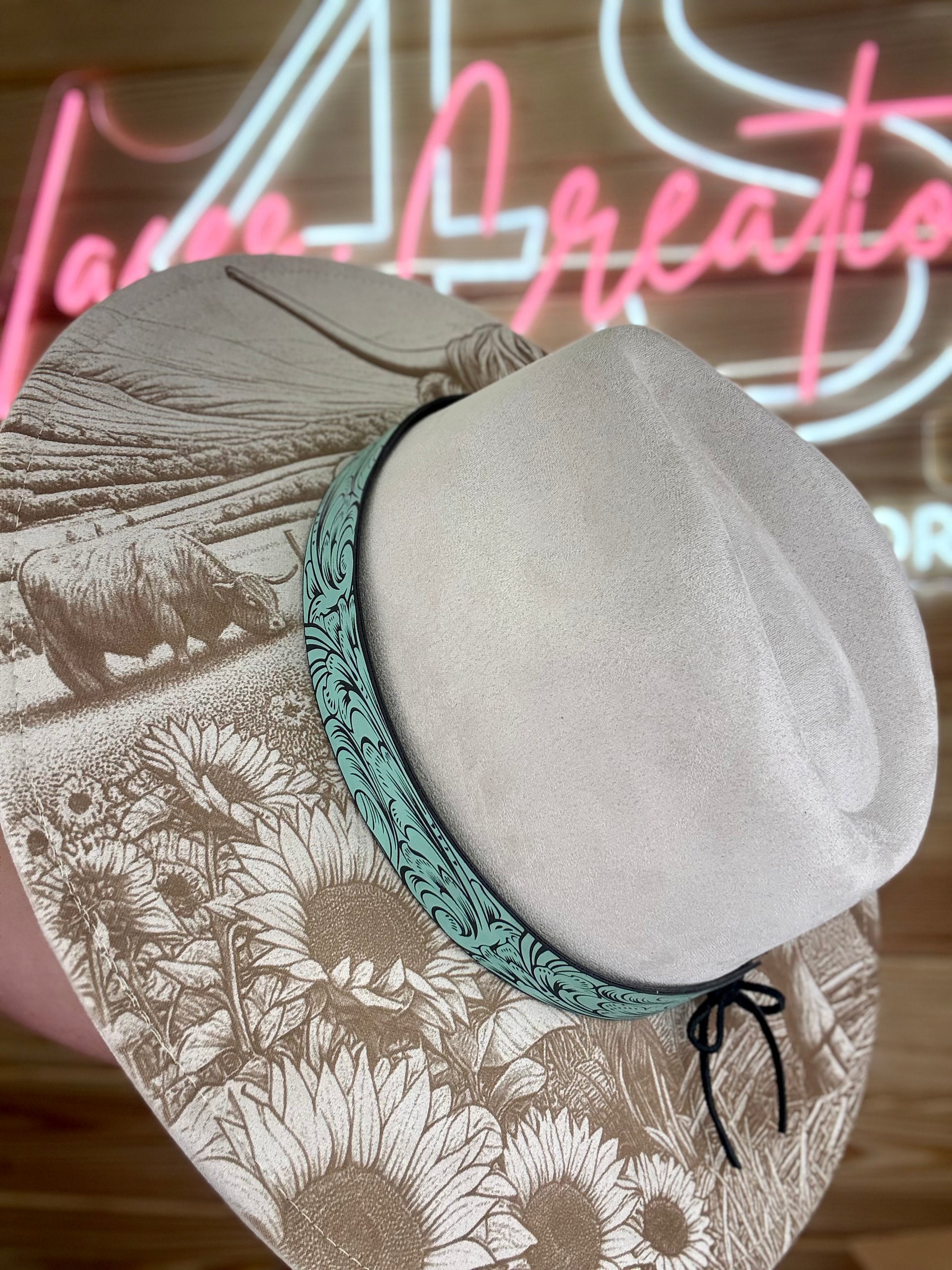 Stone colored wide brim hat with teal laser engraved head band featuring a sunflower and highland cow design