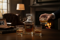 Person holding a glass of whiskey next to a decanter and glasses on a table with a warm, cozy background.