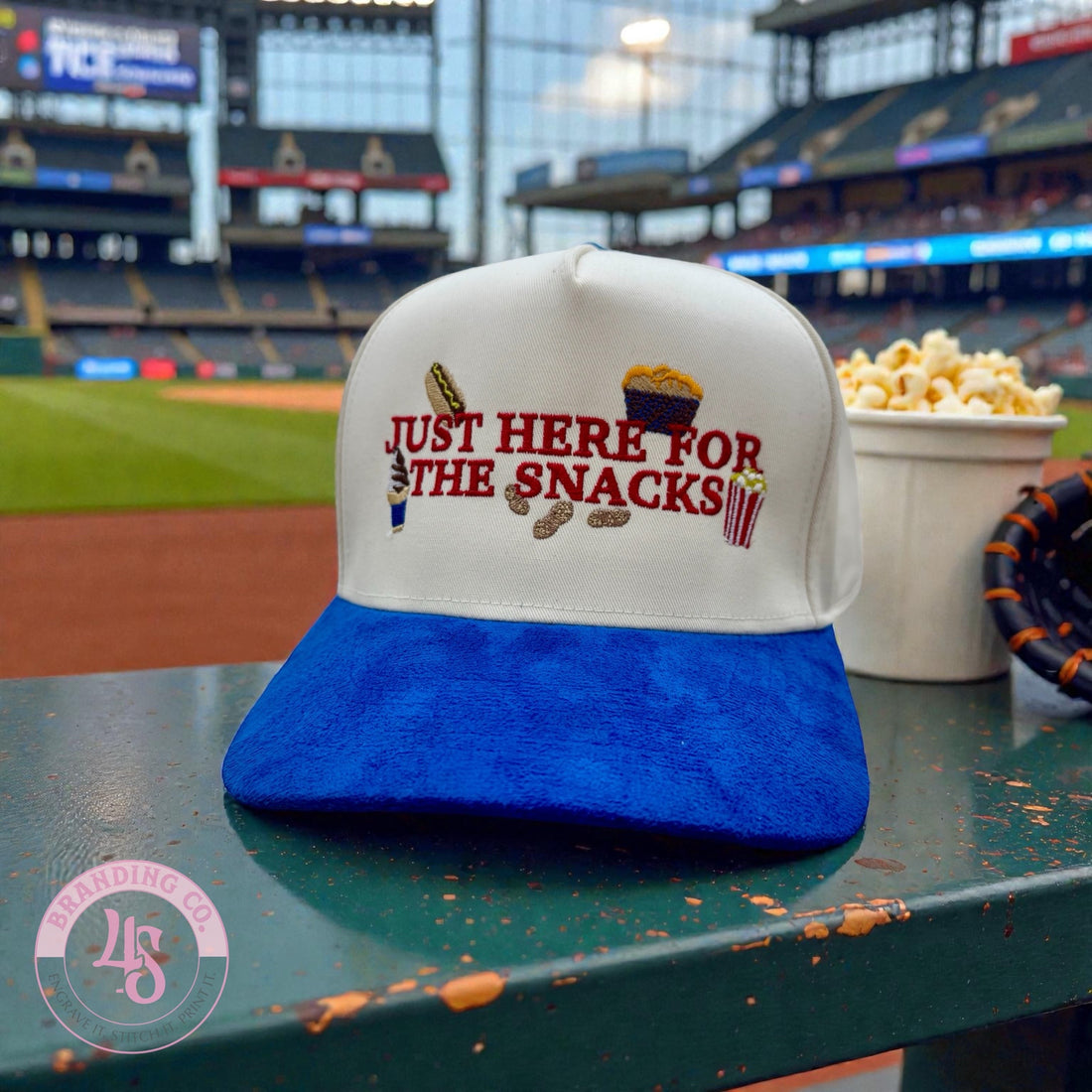 Baseball cap with 'Just Here for the Snacks' text on a table at a baseball stadium.