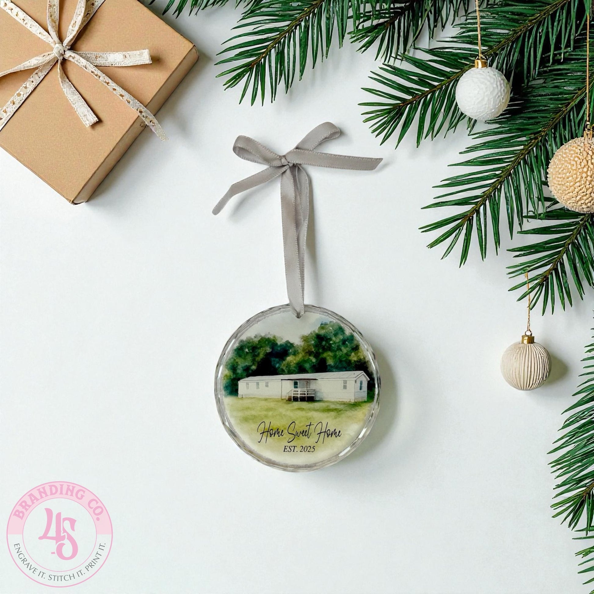 Round Christmas ornament with a house illustration and 'Home Sweet Home' text, tied with a ribbon, surrounded by festive decorations on a white background.